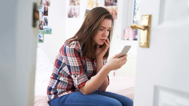 Foto: Shutterstock. Jente ser på mobilen sin på rommet sitt, litt sjokkert.