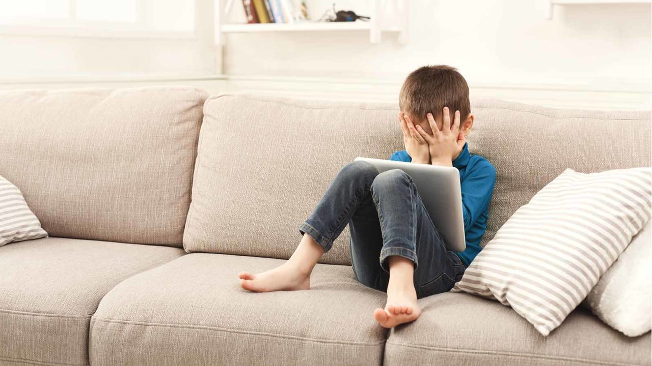Gutt sitter i sofa, holder seg for øynene, har et nettbrett i fanget. Foto: Shutterstock.