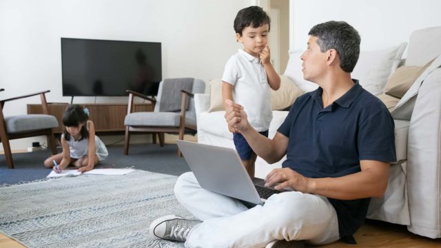 Foto: Shutterstock. Bildet viser en pappa sittende på gulvet i stuen med en datamaskin. Sønnen står ved siden av. De snakker sammen.