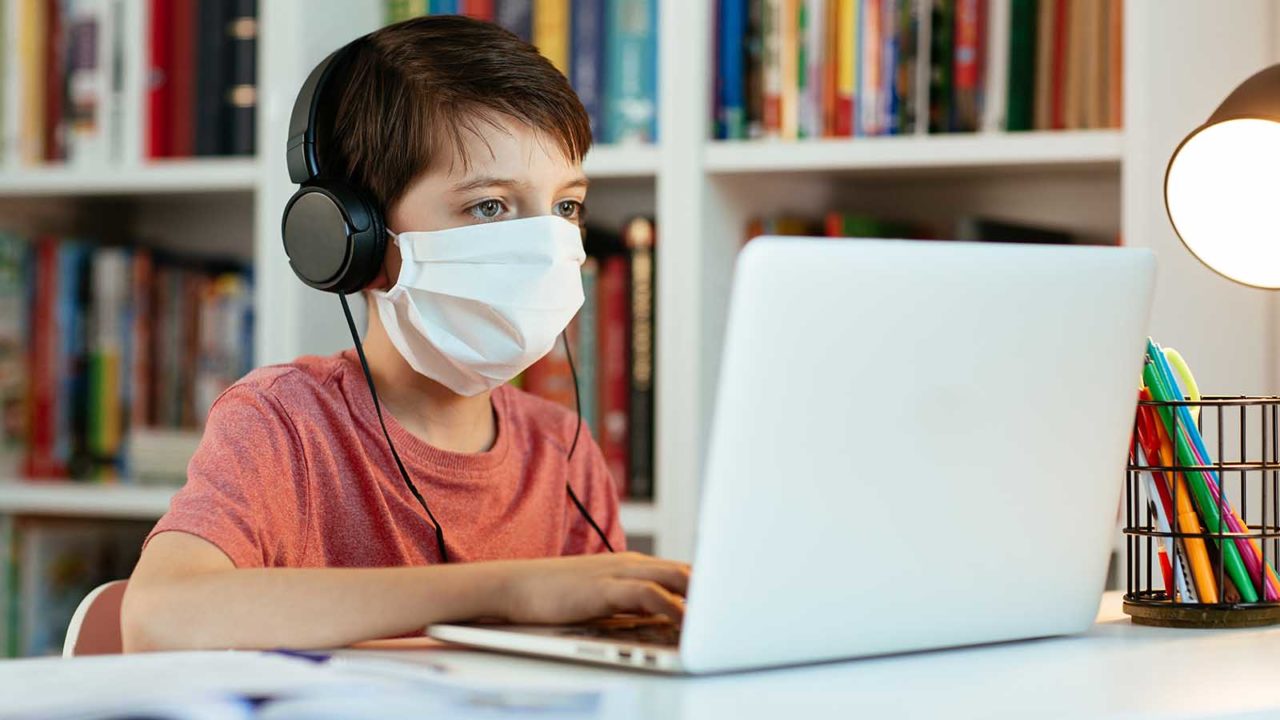 Foto: Shutterstock. Bildet viser en gutt med medisinsk munnbind Han har hodetelefoner og er på nett.