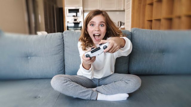 Foto: Shutterstock. Bildet viser ei jente med spillkontroller. Hun sitter i en sofa i en stue og er svært engasjert i spillingen.
