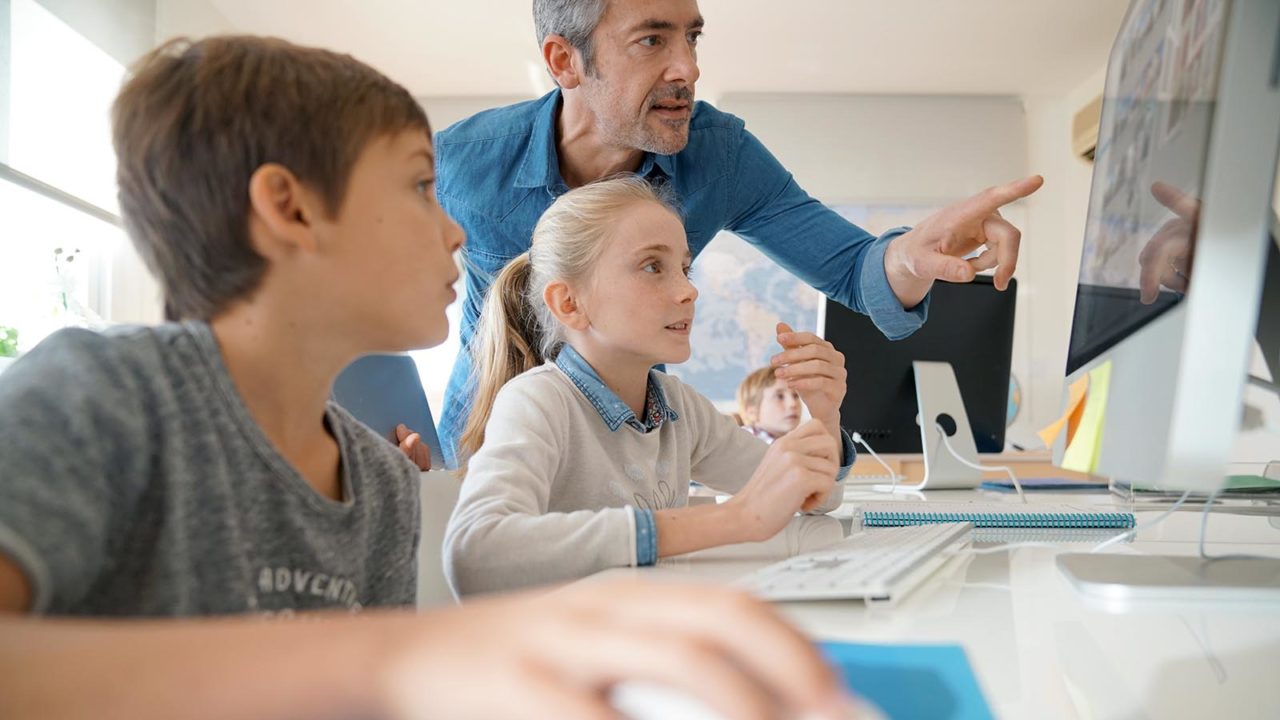 Foto: Shutterstock. Bildet viser to elever med datamaskiner og en lærer som peker på skjermen.