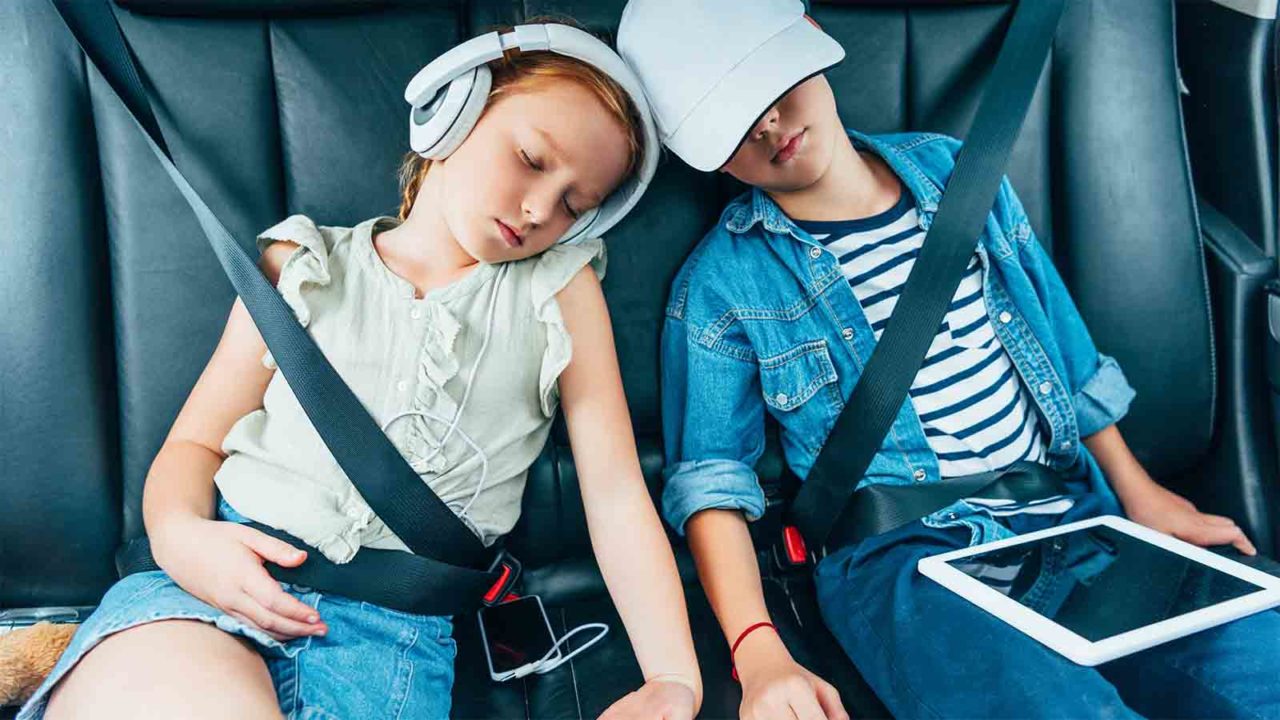 Foto: Shutterstock / LightField Studios. Bildet viser to barn som har sovnet i baksetet på en bil, men nettbrett i fanget.