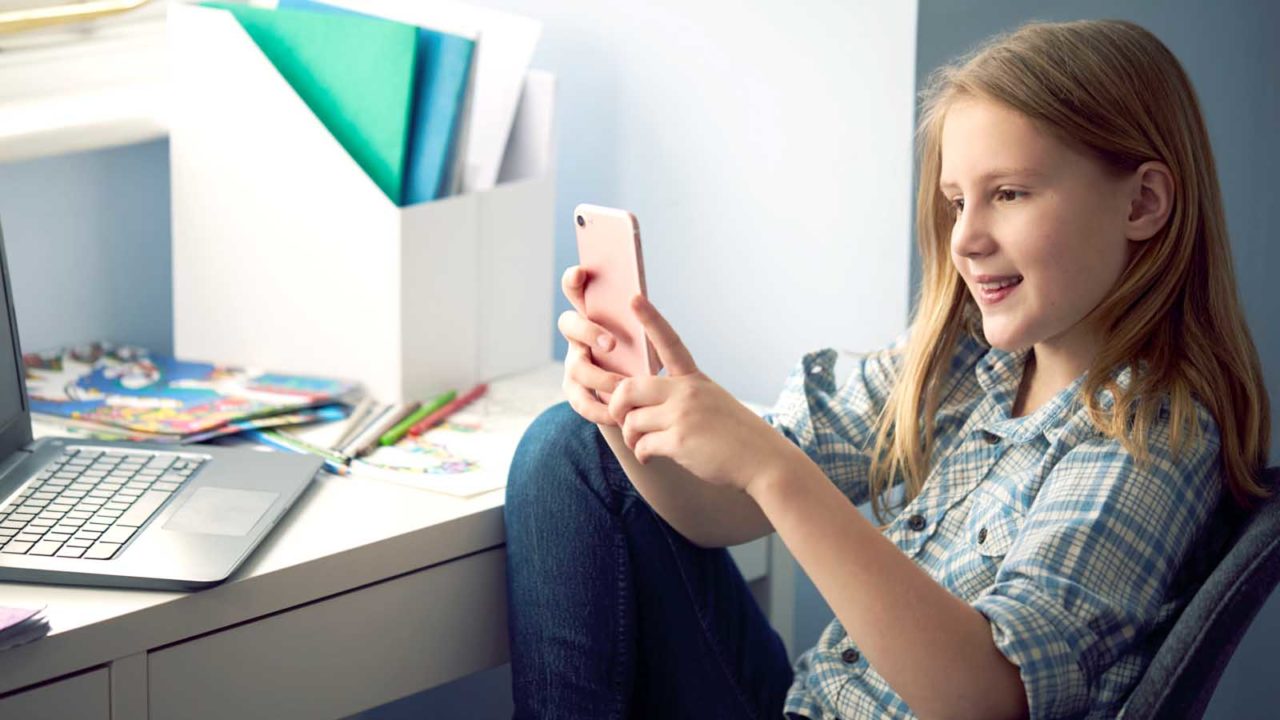 Foto: Shutterstock / Monkey Business Images. Bildet viser ei tenåring ved pulten sin hjemme. Hun har PC og blyanter på bordet. I hånden holder hun en telefon.