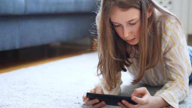 Foto: Shutterstock / M-Production. Bildet viser ei jente som ligger på gulvet og ser på en mobiltelefon.