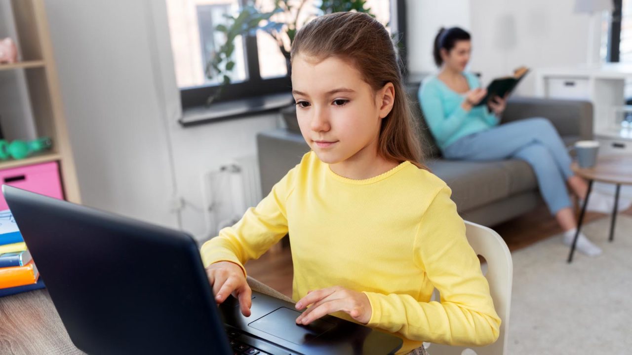 Foto: Shutterstock / Syda Productions. Bildet viser ei jente som skriver på tastaturet til en datamaskin hjemme i stua, moren sitter og leser en bok i sofaen i bakgrunnen.