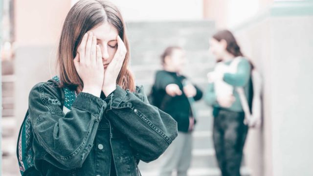 Foto: Shutterstock / Melinda Nagy. Bildet viser en fortvilet tenåringsjente som holder hendene foran ansiktet. To jenter stå i bakgrunnen og gjør narr av henne.