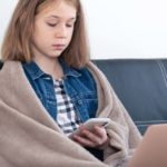 Foto: Shutterstock / PV productions. Bildet viser ei jente i sofaen, med teppe rundt seg, hun ser på telefon og har datamaskin på fanget.