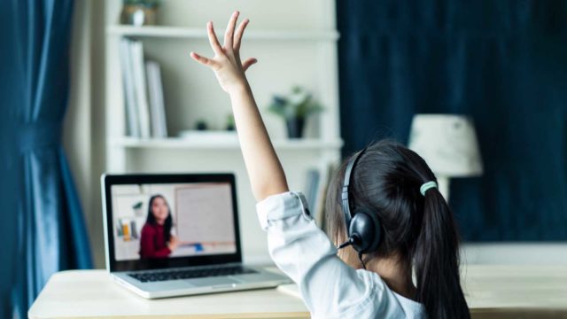 Foto: Shutterstock / Hananeko_Studio. Bildet viser ei jente som rekker opp hånden. Hun sitter hjemme og følger undervisning på skjerm.