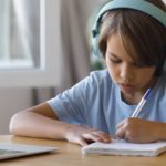 Foto: Shutterstock / fizkes. Bildet viser en gutt med hodetelefoner og datamaskin, han noterer på ark med kulepenn.