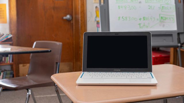 Foto: Shutterstock / CC Photo Labs. Bildet viser et klasserom med en tustavle og noen pulter. På pulten stpr en oppslått datamaskin av typen Chromebook.