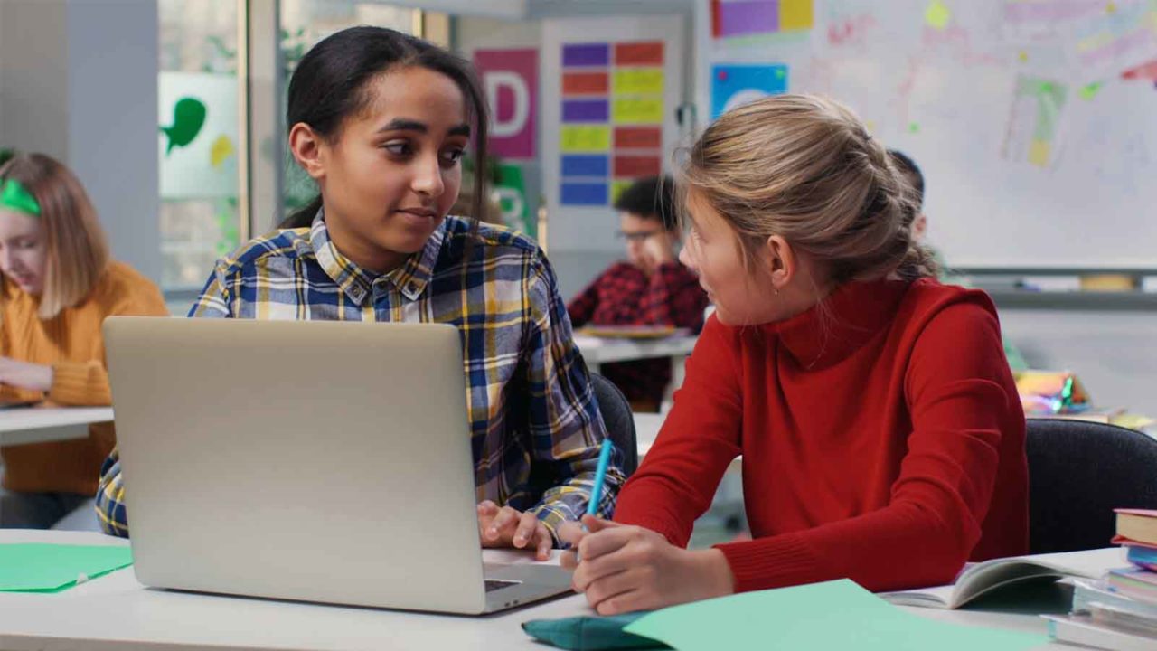 Foto: Shutterstock / nimito. Bildet viser to jenter som er elever, i et klasserom, med en datamaskin på pulten.