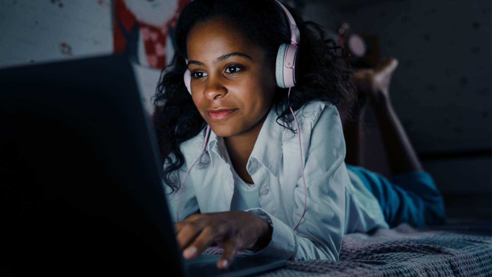 Foto: Shutterstock / Dean Drobot. Bildet viser ei tenåringsjente liggende på sengen i et ganske mørkt rom, hunhar på seg hodetelfoner og skriver på en datamaskin..