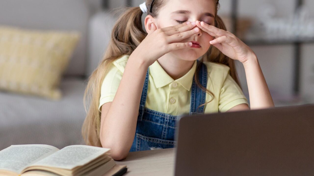 Foto: Shutterstock / Prostock-studio. Bildet viser ei jente som gnir seg i øynene og ser trøtt ut, foran på bordet er en PC og en bok.