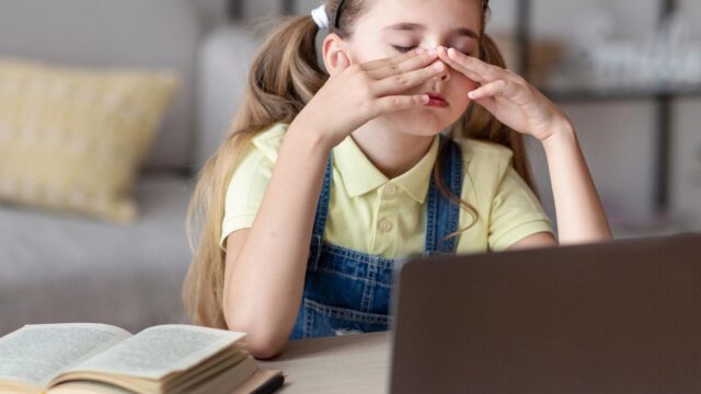 Foto: Shutterstock / Prostock-studio. Bildet viser ei jente som gnir seg i øynene og ser trøtt ut, foran på bordet er en PC og en bok.