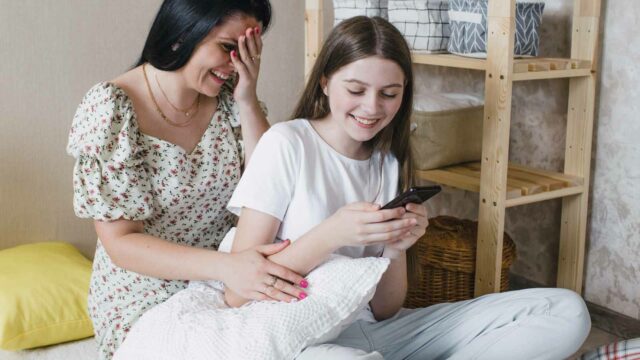 Foto: Shutterstock / Miridda. Bildet viser en mor og en datter som ler av en vits de leser på telefonen.