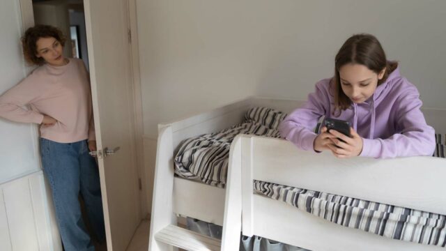 Foto: Shutterstock / fizkes. Bildet viser en bekymret mor i døråpningen inn til datterens soverom. Datteren skriver på telefonen.