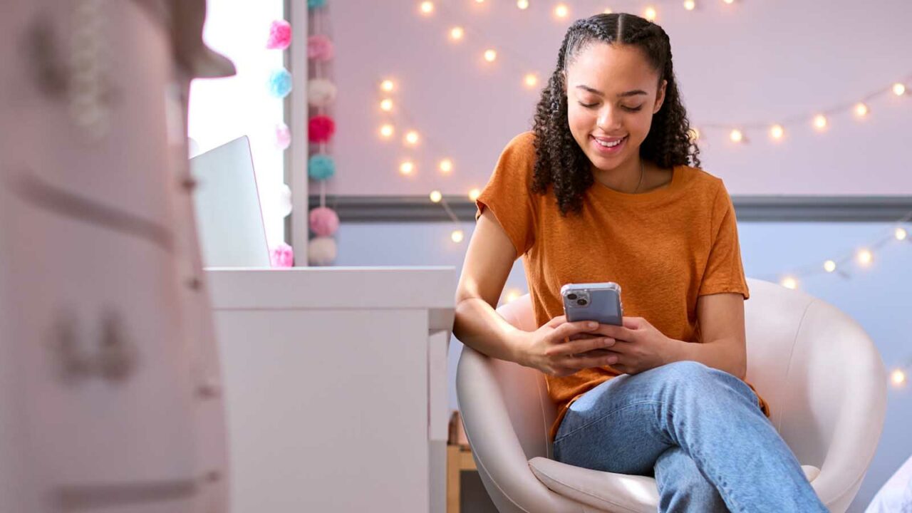 Foto: Shutterstock / Monkey Business Images. Bildet viser ei tenåringsjente som sitter i en stol på rommet sitt og leser noe på mobiltelefonen.