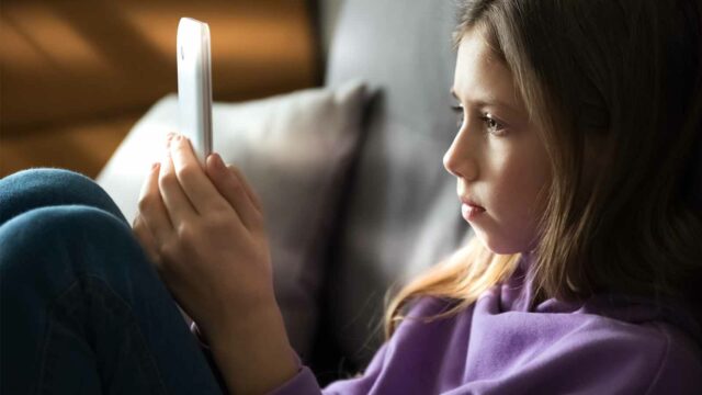 Foto: Shutterstock / Iren_Geo. Bildet viser ei alvorlig jente hjemme som ser på skjermen på en telefon.
