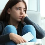 Foto: Shutterstock / New Africa. Bildet viser ei jente som ser trist og tankefull ut. Hun sitter i en sofa og holder en telefon i hånden.