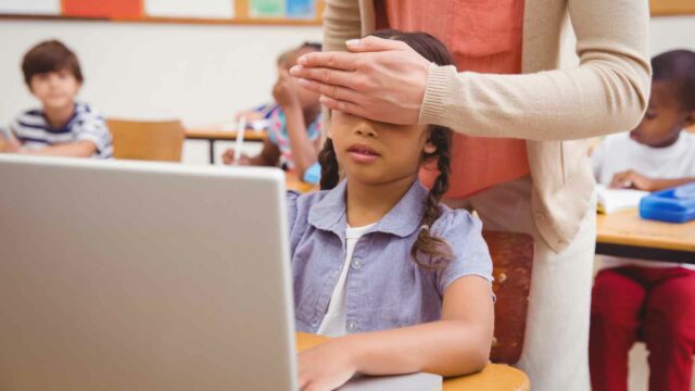 Lærer holder hånden foran øynene på elev som sitter med datamaskin på pulten. Foto: Shutterstock / wavebreakmedia.